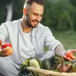 Man seated outdoors with a basket of fresh vegetables, enjoying the garden.
