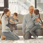 Happy parents with children on knees in casual clothes sitting on floor and playing with kids while spending time together in living room near panoramic window