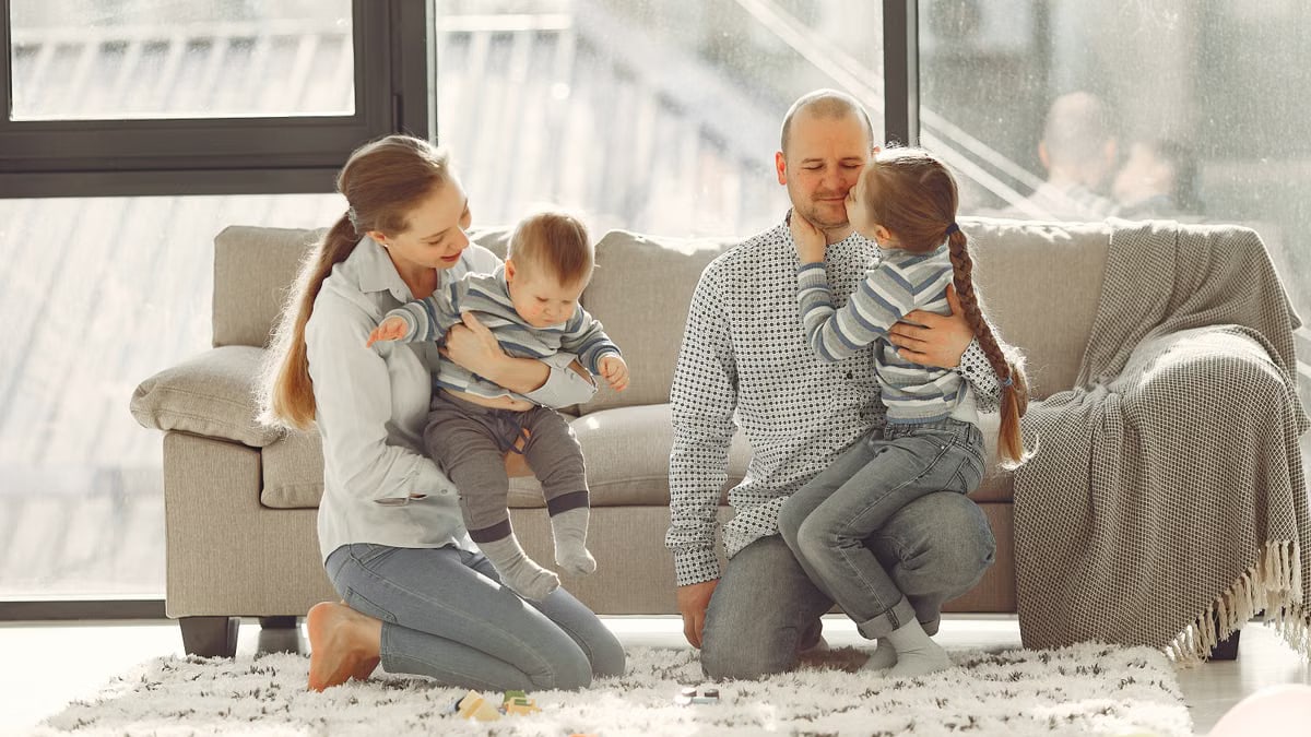 Happy parents with children on knees in casual clothes sitting on floor and playing with kids while spending time together in living room near panoramic window